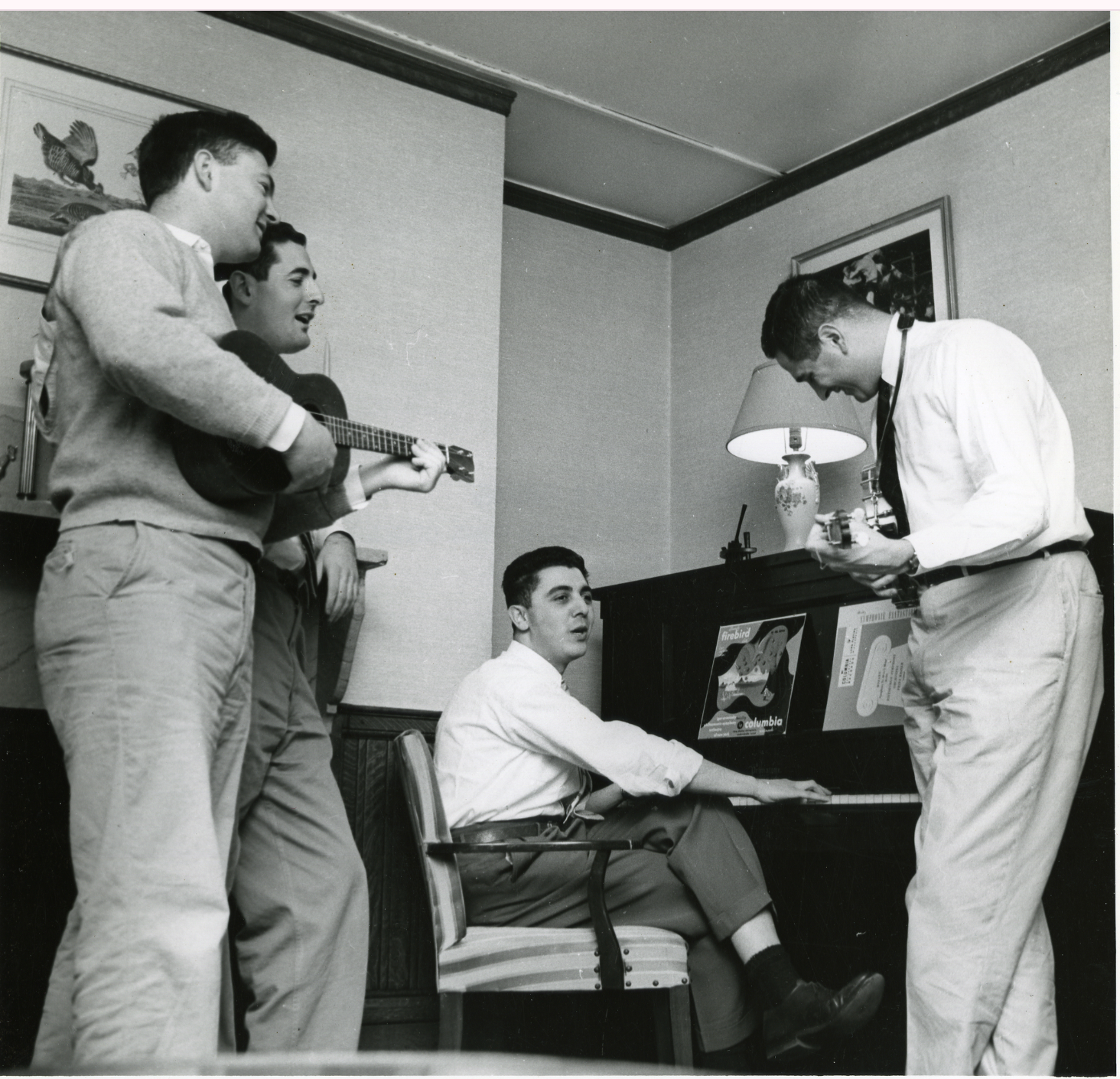 All most full-length view of three men standing around another man who is sitting at an upright piano. The man standing closest to the camera on the left is holding a ukulele.